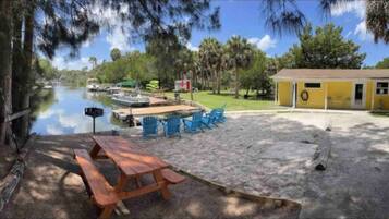 A sandy area with a picnic table and blue chairs, a small yellow building, and a calm water body with boats.