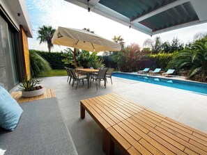 Un patio exterior con piscina, una mesa de madera y sillas bajo un toldo.
