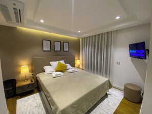 Quarto de hotel moderno com cama bem arrumada, televisão de tela plana, mesa de cabeceira com abajur e ar condicionado na parede.