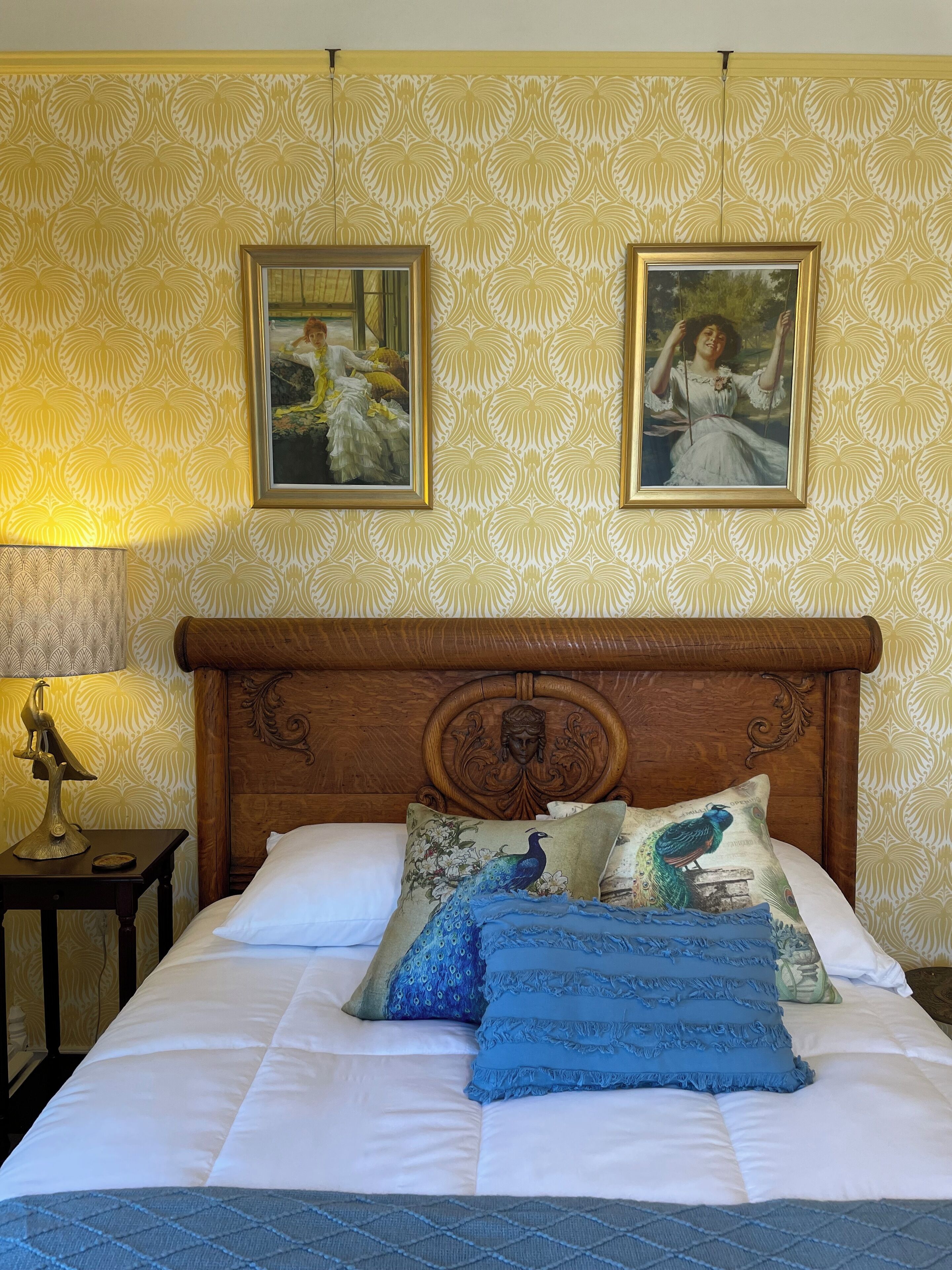 Una cama bien hecha con sábanas blancas y almohadas decorativas, cabecera de madera con un diseño ornamentado y dos cuadros en la pared.