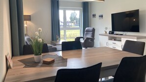 A modern living room with a dining area, a flat-screen TV, and a large window with curtains.