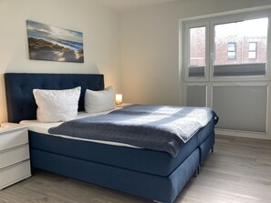 Un dormitorio con cama, mesita de noche, lámpara, ventana con persianas y un cuadro en la pared.