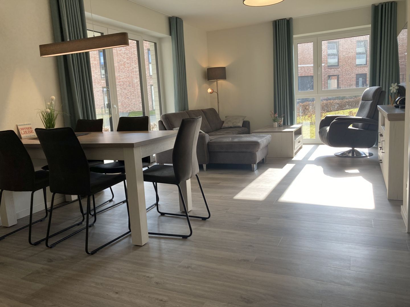 Un salón moderno con comedor, ventanas amplias y vista a un edificio de ladrillo.