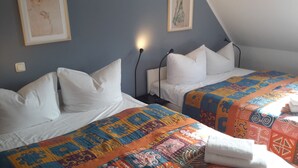 Dos camas individuales con sábanas blancas y colchas coloridas, una mesita de noche con una lámpara y un cuadro en la pared.