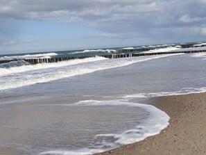A beach with waves crashing onto the shore, a wooden pier extending into the water, and a lighthouse in the distance. - Beach view 100m (Kühlungsborn)