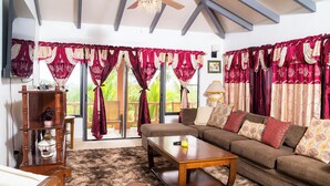 A living room with a sofa, coffee table, and wooden furniture. There are curtains, a chandelier, and a window with a view.