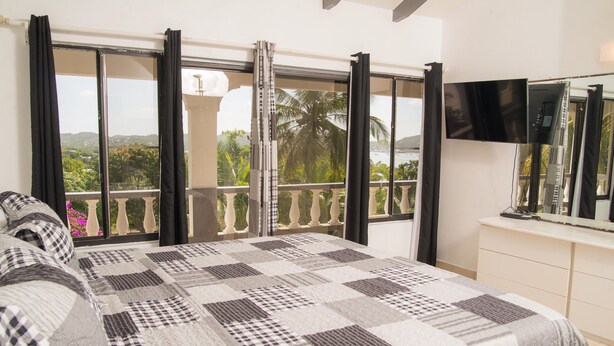 A bedroom with a checkered bedspread, a balcony with curtains, a TV on the wall, and a view of greenery outside.
