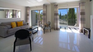 Ein geräumiges Wohnzimmer mit grauem Sofa, schwarzem Sessel und Couchtisch. Ein großes Fenster mit Vorhängen, ein Balkon mit Geländer und Blick auf Bäume und einen blauen Himmel.