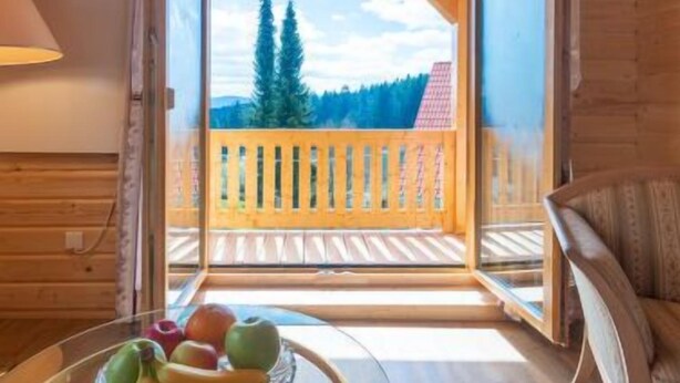 A wooden cabin interior with a balcony, a round glass table with fruit, and a view of trees and mountains.