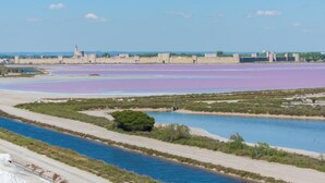 Paisagem costeira com rio, salinas e uma fortificação histórica.