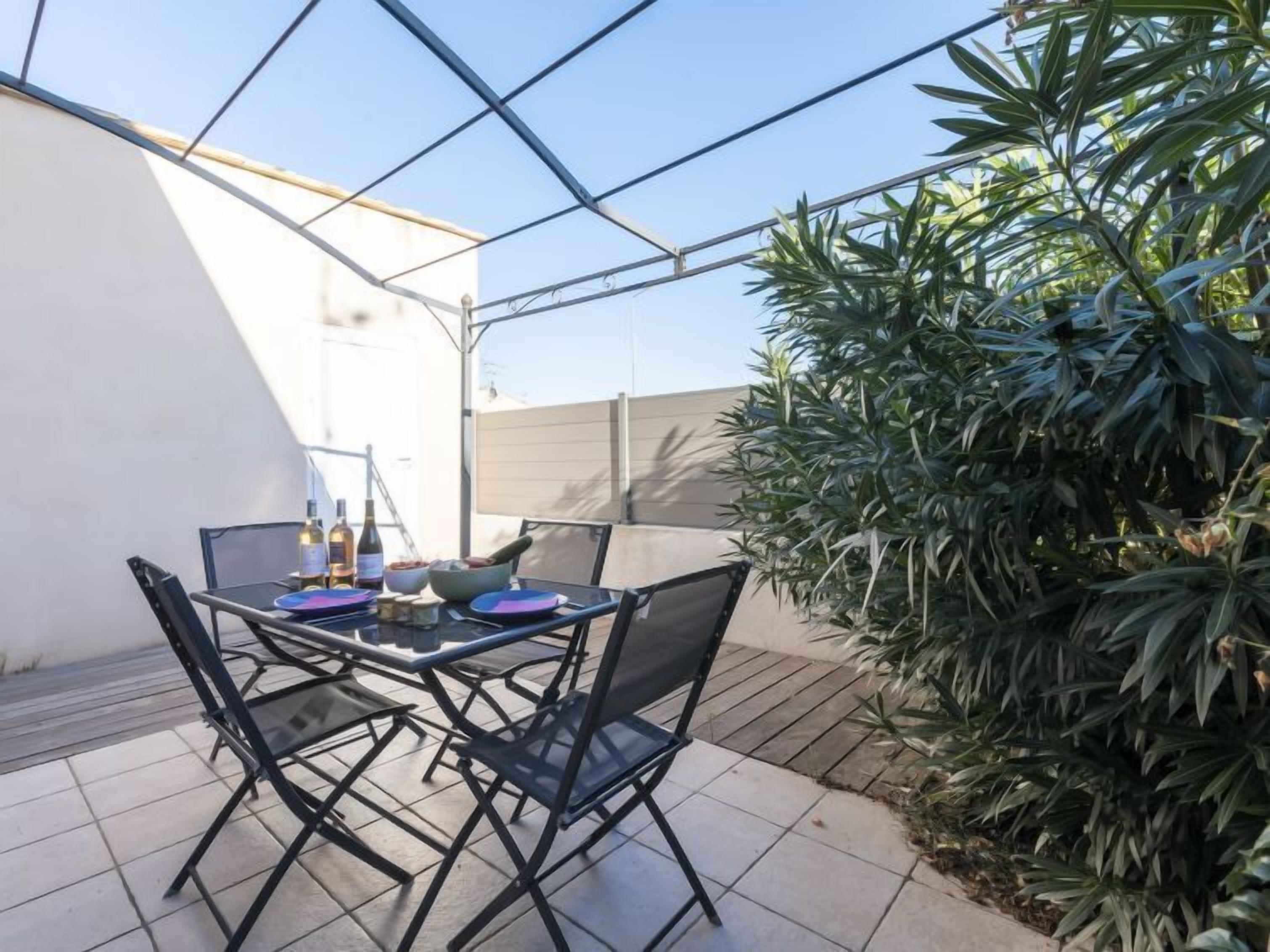 Un patio exterior con una mesa y sillas, rodeado de plantas y una pared blanca.