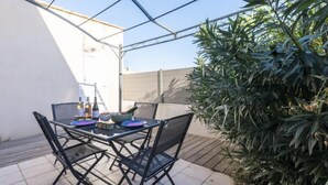 Um pátio externo com mesa e cadeiras, rodeado por plantas e uma parede branca.