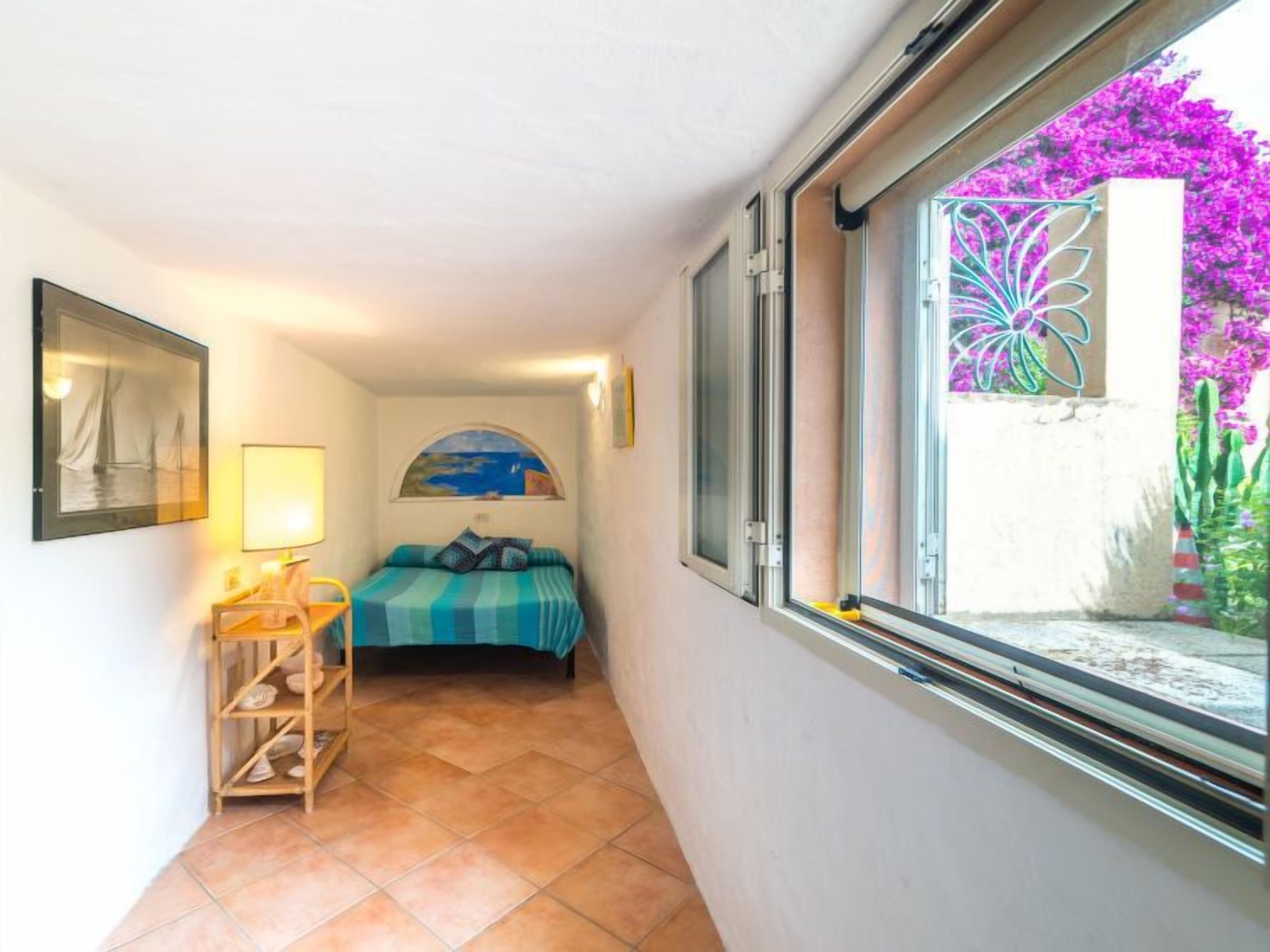 Un pasillo estrecho con una cama, un estante con toallas, un cuadro enmarcado y una ventana que da a un jardín.