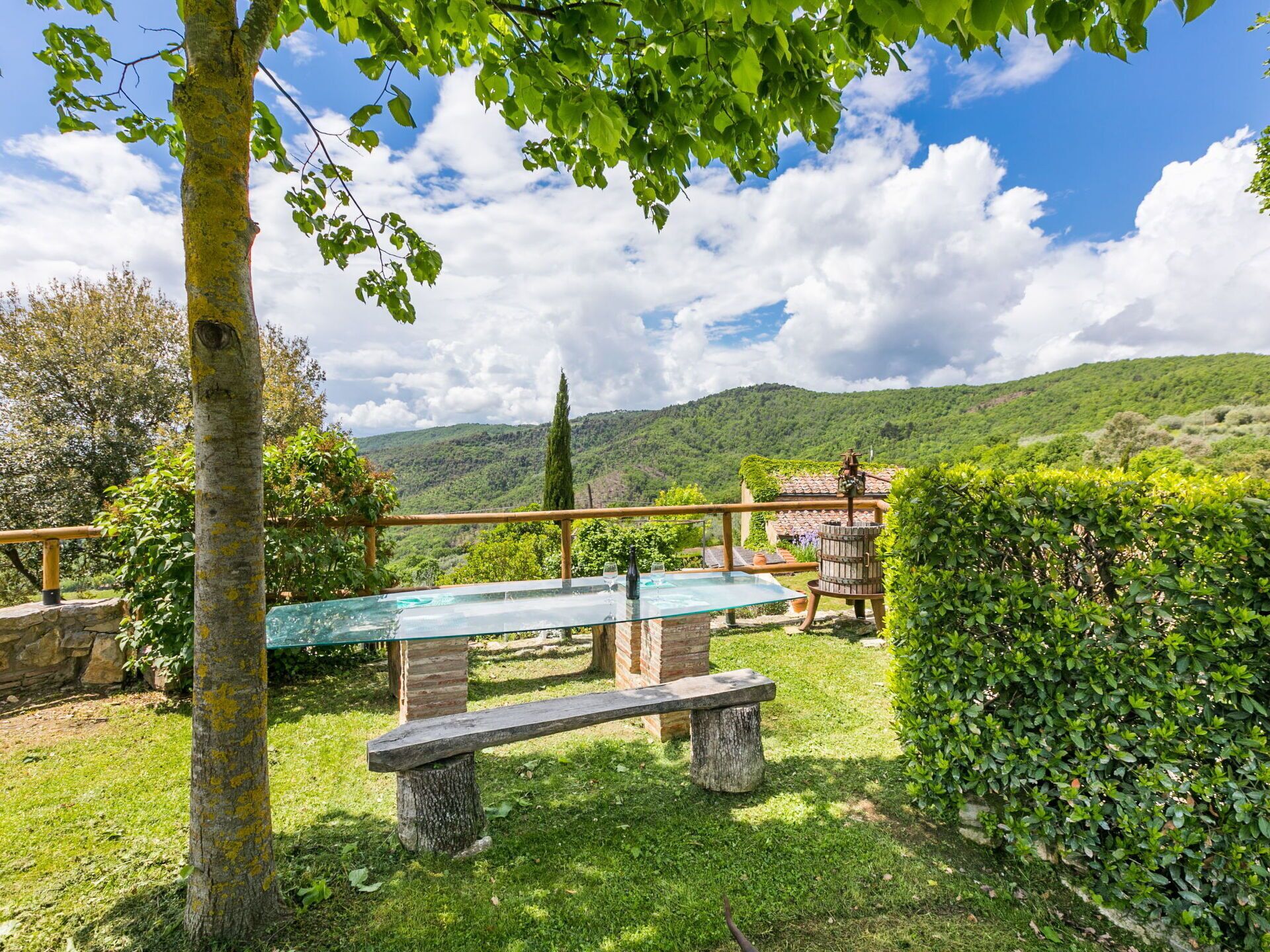 A glass-top table with a bench, surrounded by greenery and a view of rolling hills.