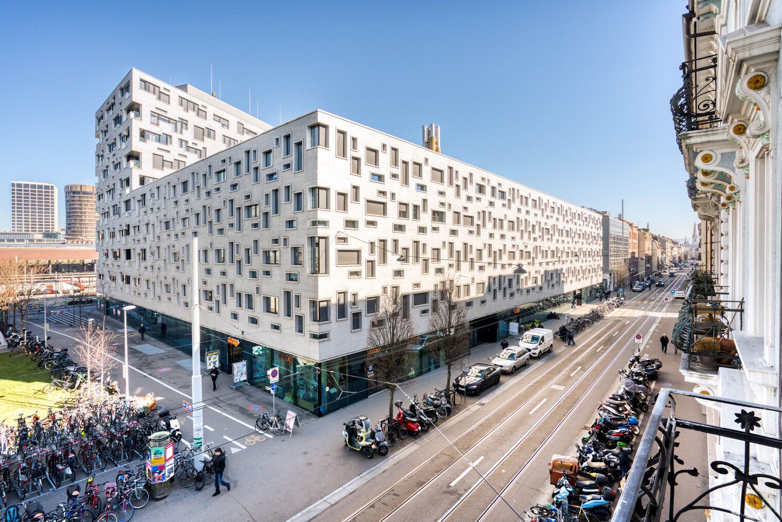 Ein modernes Gebäude mit einer rasterförmigen Fassade, einem Ladenbereich im Erdgeschoss und einer Straßenbahn, die daran vorbeifährt.