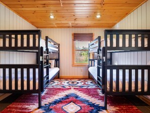 A room with bunk beds, a patterned rug, wooden walls, and a window with blinds.