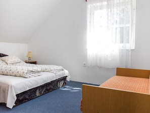 A bedroom with a bed, a nightstand, a lamp, a window with sheer curtains, and a small wooden table.