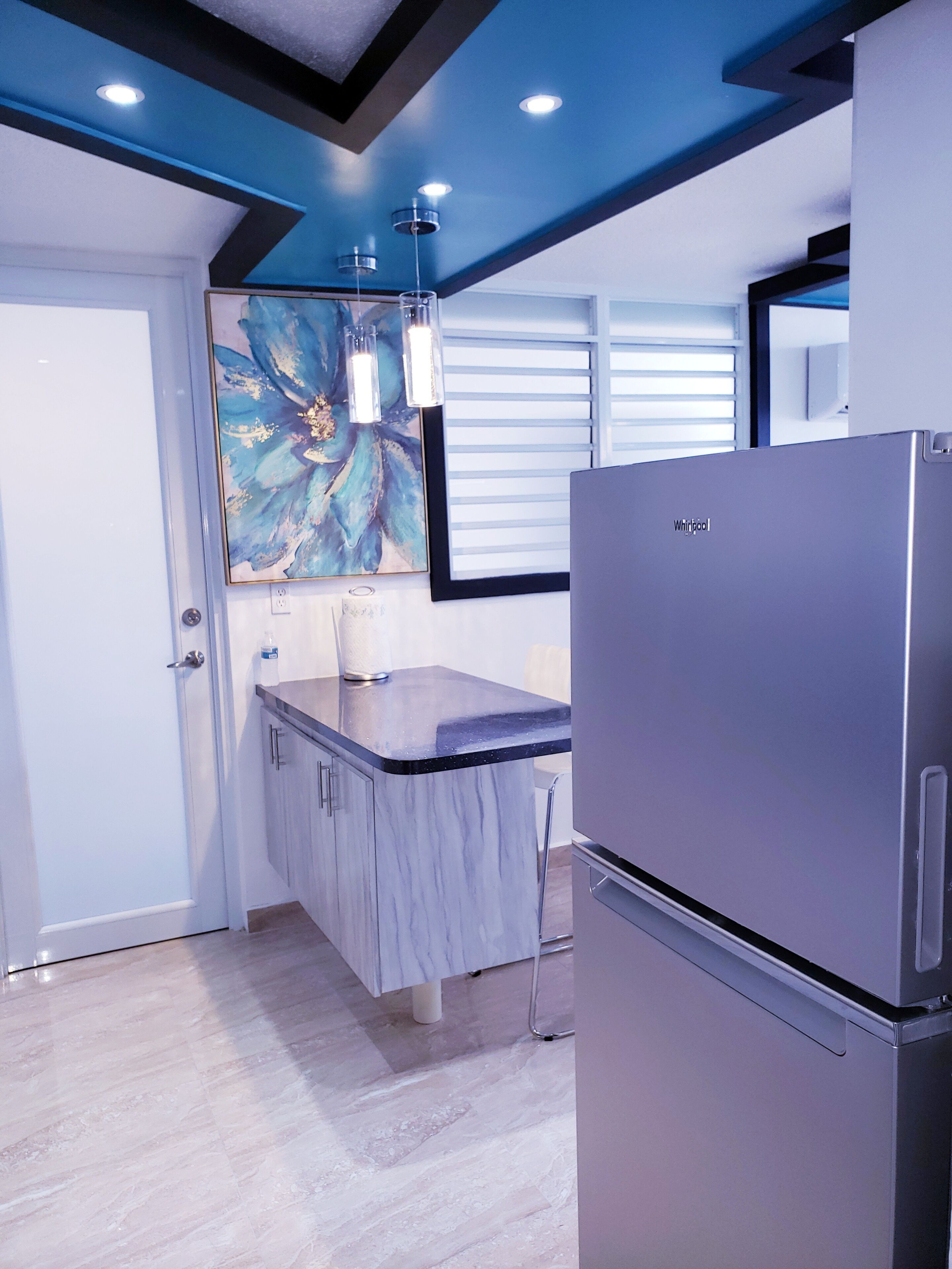 A modern kitchen with a marble countertop, a stainless steel refrigerator, and a blue ceiling with recessed lighting.