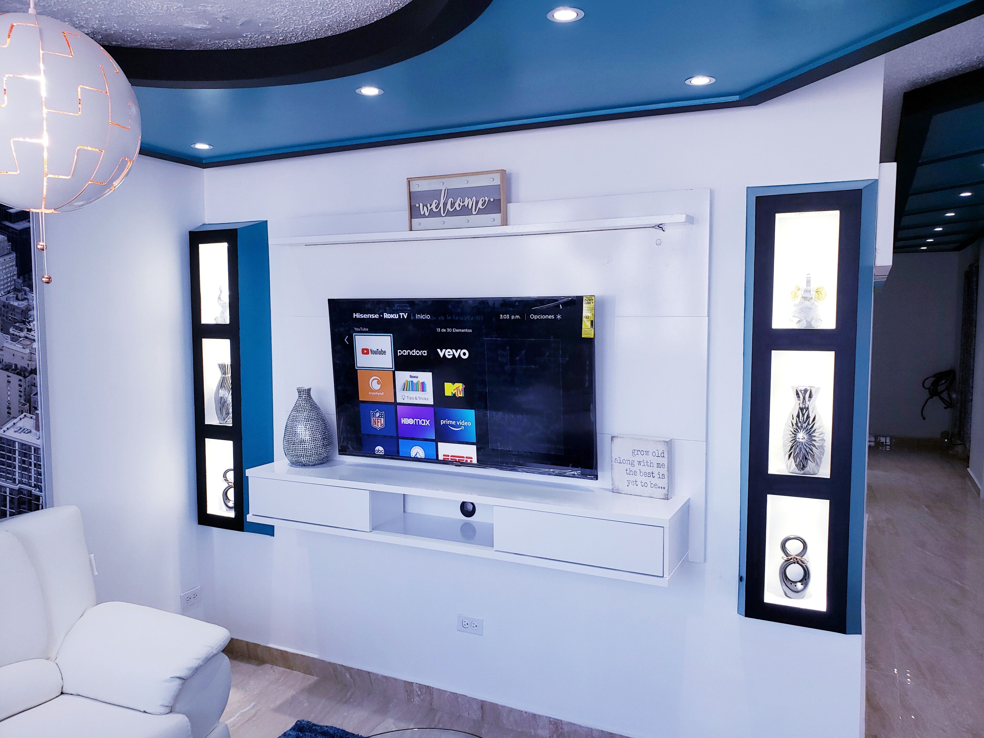 Un soggiorno moderno con una TV a parete che mostra diversi servizi di streaming, un divano bianco e un vaso decorativo.