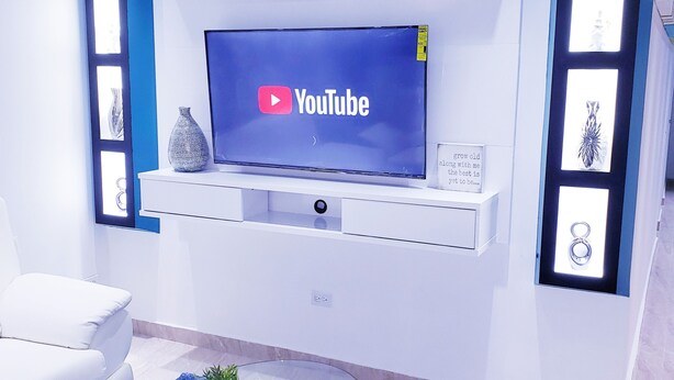 Una sala de estar moderna con un televisor de pantalla plana montado en la pared, un sofá blanco, una mesa de centro de vidrio y objetos decorativos en los estantes.