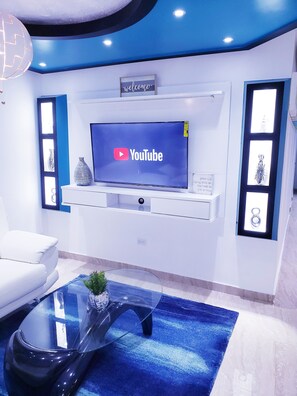 A modern living room with a flat-screen TV mounted on a wall, a white sofa, a glass coffee table, and decorative items on the shelves. - MH (#1)⭐️beautiful house near the airport and beaches (Carolina Puerto Rico)