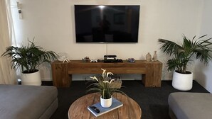 Un salon avec un téléviseur à écran plat fixé au mur, un banc en bois, deux plantes en pot, une table basse ronde avec une télécommande et un canapé.