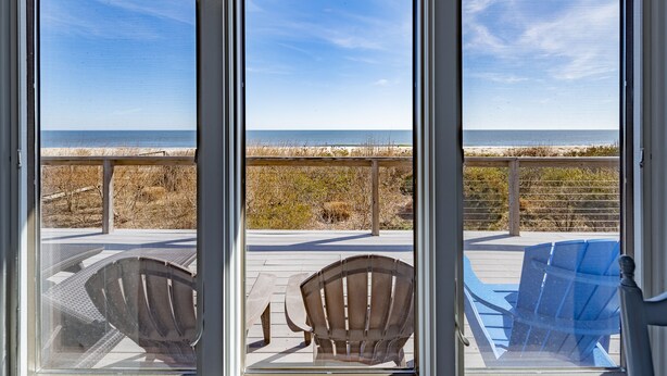 A view through a large window of a beachside property with a wooden deck, two Adirondack chairs, and a blue chair. - 6 Bedroom Oceanfront House in N. Bethany Beach. Renovated in 2024. Watch video! (Bethany Beach)