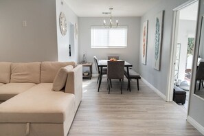A modern living room with a beige sectional sofa, a dining area with a table and chairs, and a chandelier.