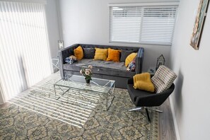 Ein Wohnzimmer mit grauem Sofa, gelben Kissen, einem Glastisch und einem gemusterten Teppich.