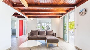 Un salon moderne avec un plafond en bois, un canapé d’angle, une porte vitrée donnant sur l’extérieur et une table blanche.