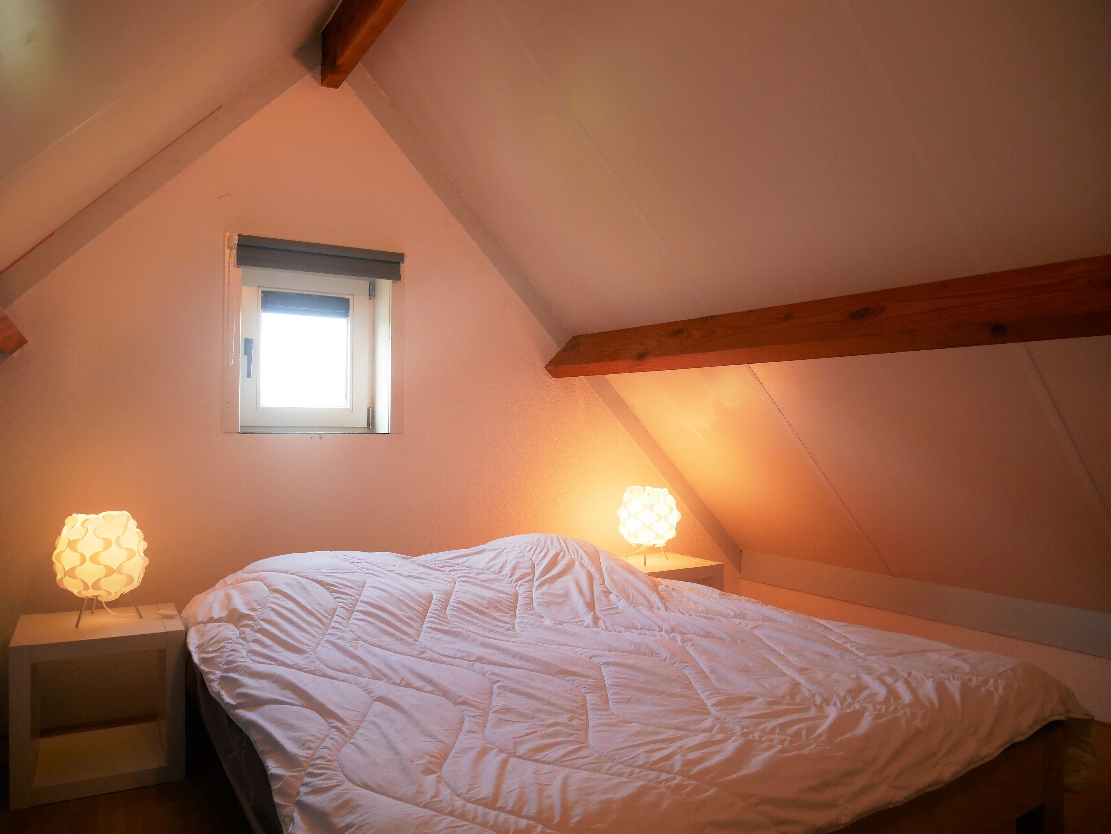 Ein Schlafzimmer mit schrägen Wänden, einem Einzelbett, zwei Nachttischlampen und einem kleinen Fenster.
