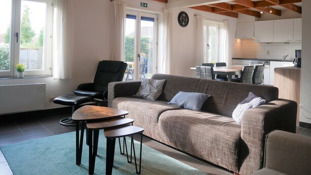 A cozy living area with a sofa, chairs, and a coffee table. There is a kitchen in the background with cabinets and a sink.