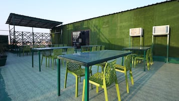An outdoor seating area with tables and chairs, a covered section, and air conditioning units on a green wall.