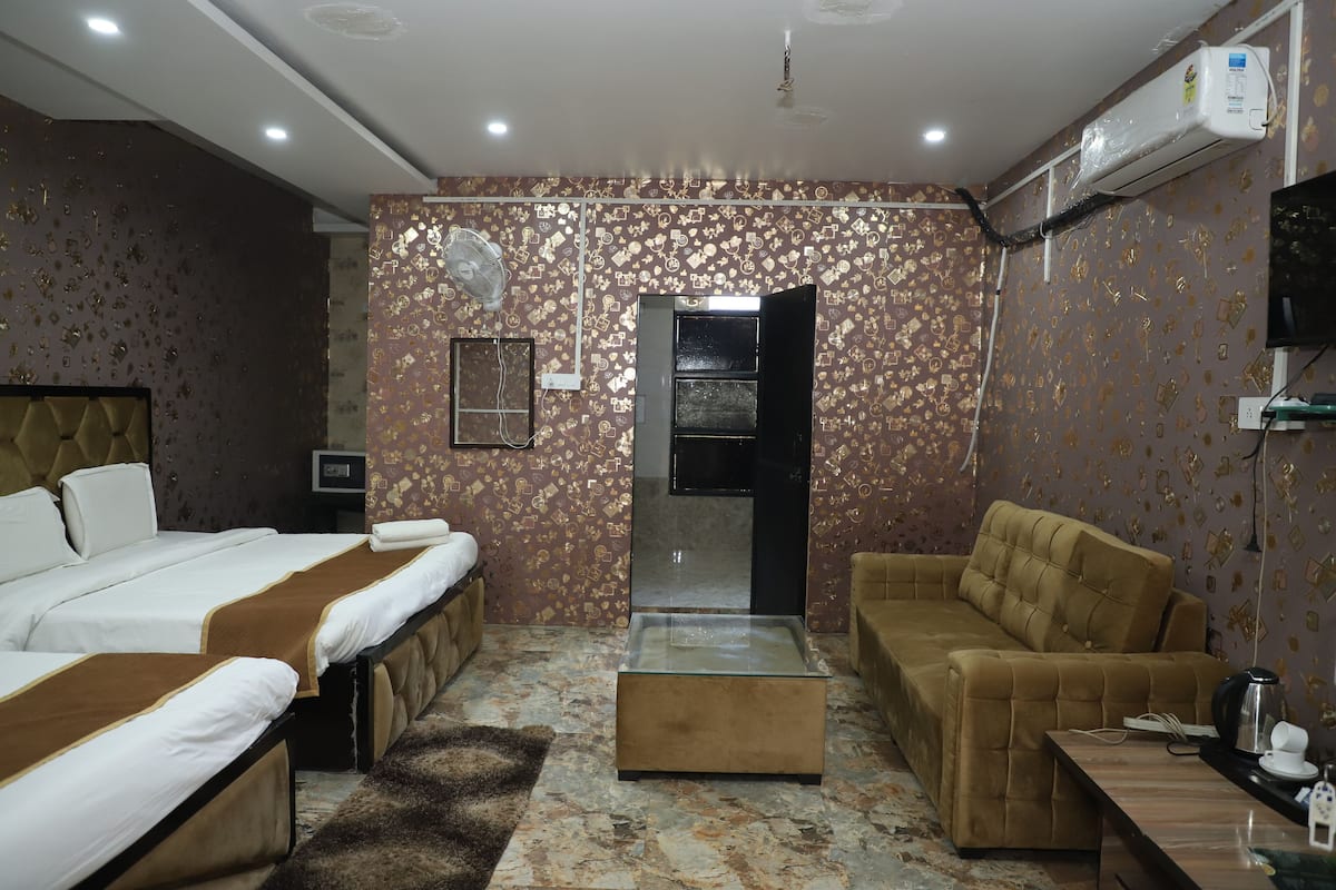 A hotel room with two beds, a sofa, a glass-top coffee table, a TV, and an air conditioning unit.