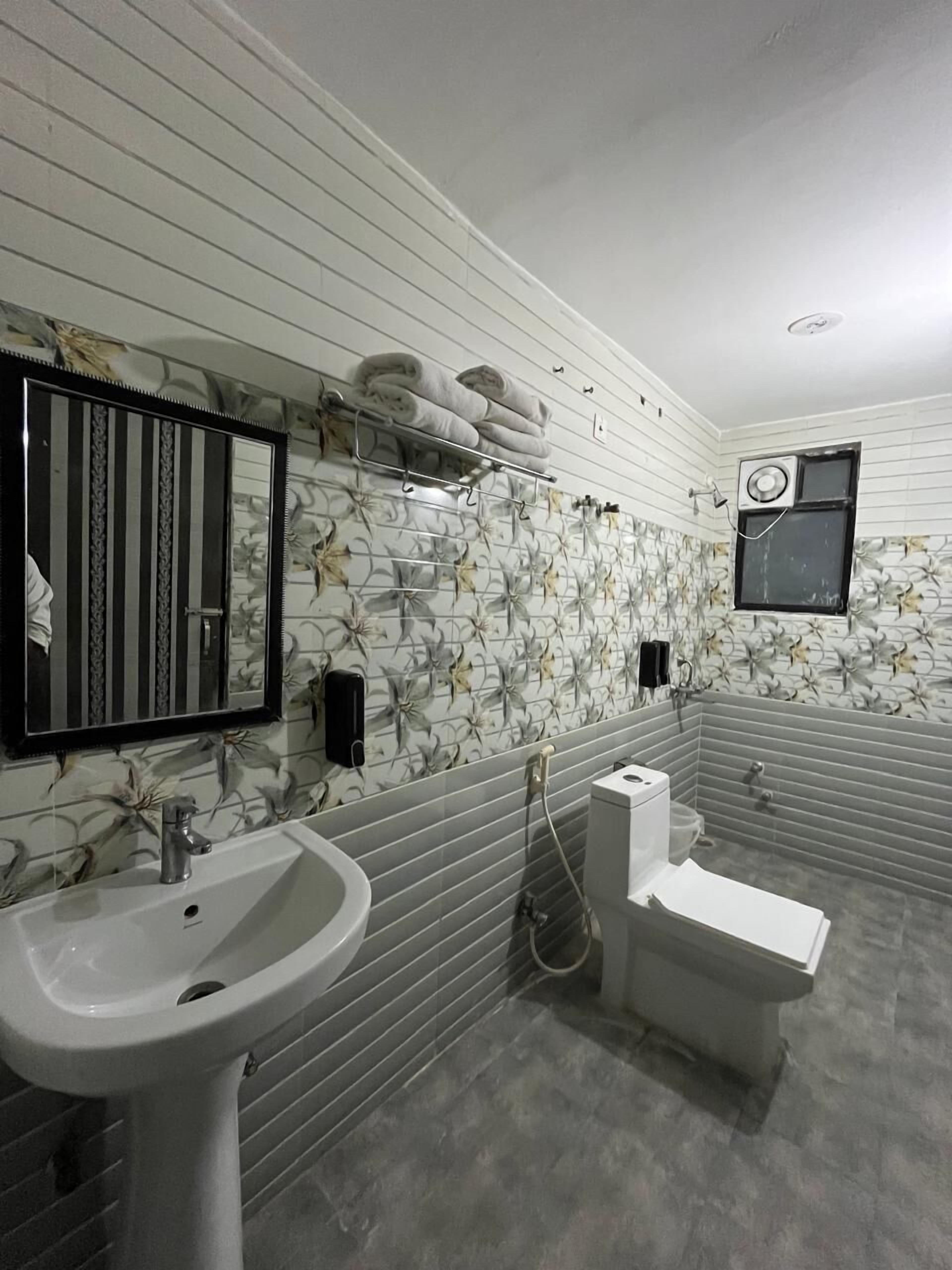 A bathroom with a white sink, a toilet, and a shower area. There is a mirror with a black frame and a towel rack with towels. The walls have a floral tile pattern.