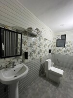 A bathroom with a white sink, a toilet, and a shower area. There is a mirror with a black frame and a towel rack with towels. The walls have a floral tile pattern.