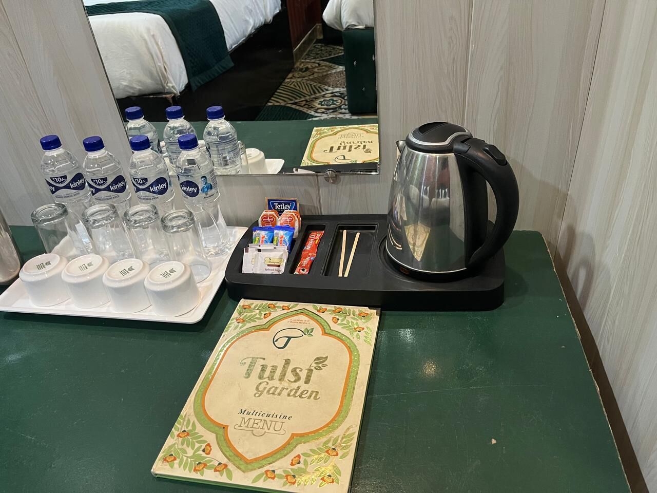Habitación de hotel con una mesa verde que tiene un tray con agua embotellada, una tetera y un menú del Tulsi Garden.
