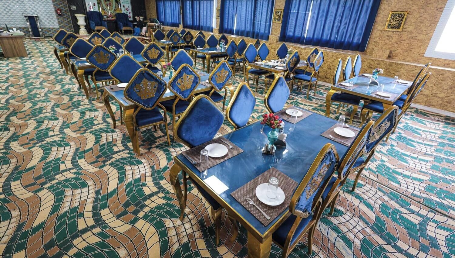 A banquet hall with rows of tables and chairs, a patterned floor, and blue curtains.