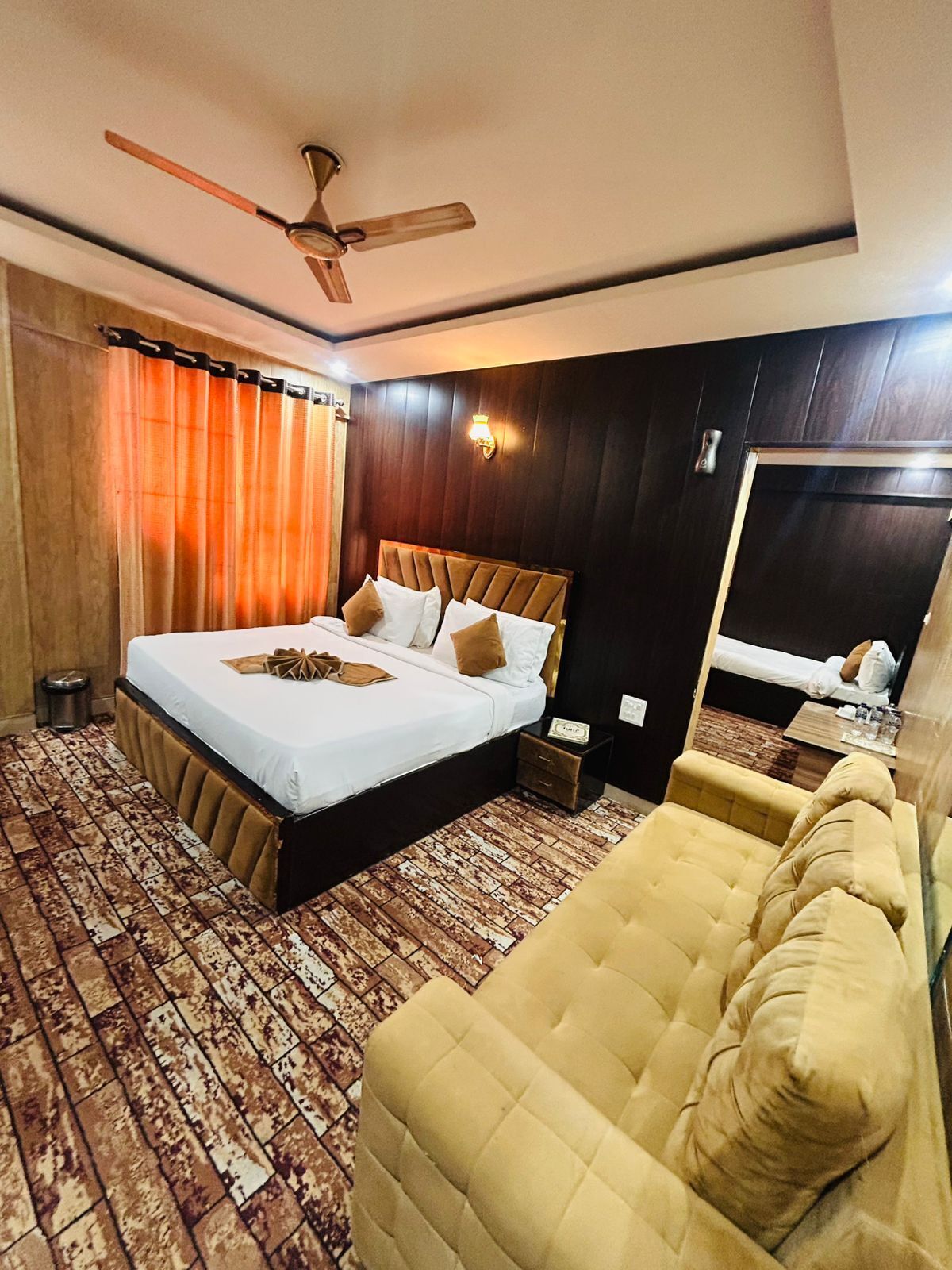 Une chambre d’hôtel avec un lit, un canapé, un ventilateur de plafond, un miroir et un plancher en bois.