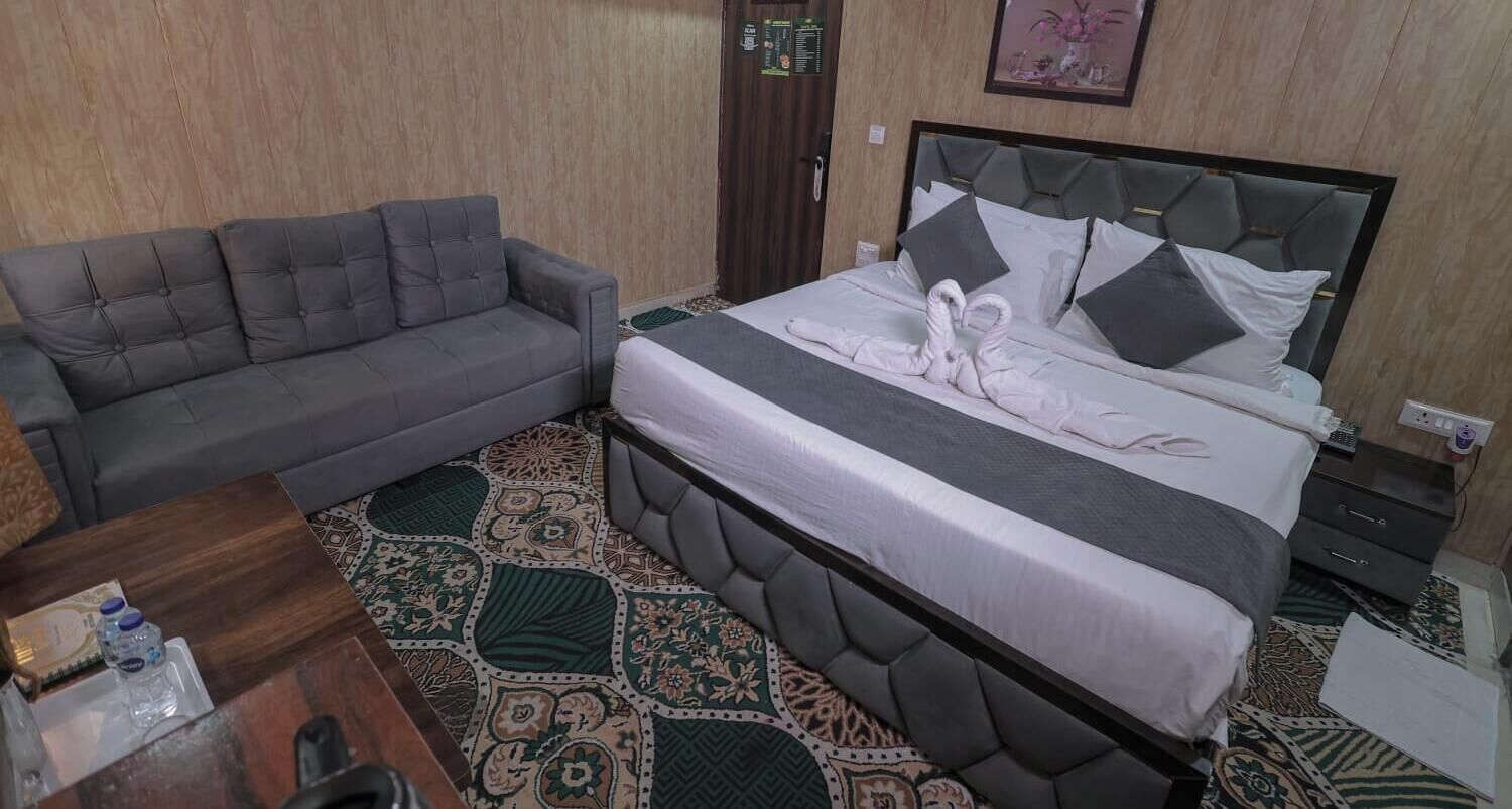 A hotel room with a grey sofa, a bed with a grey headboard, a patterned rug, and a small bedside table.