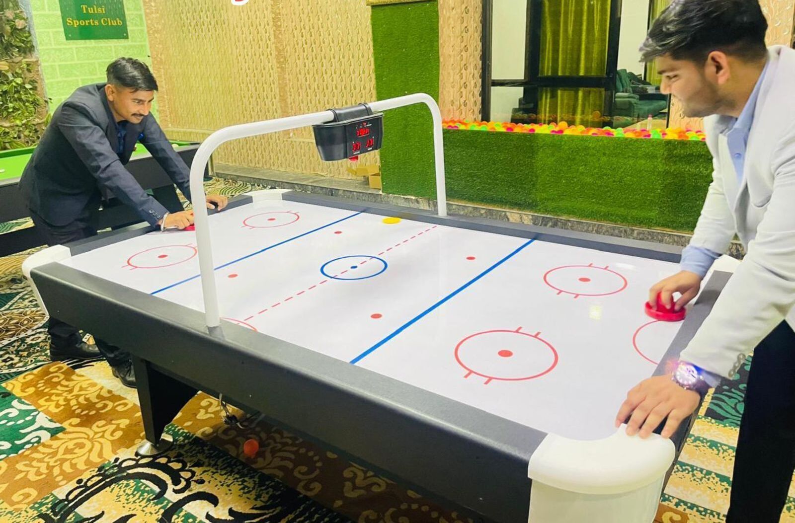 An indoor ice hockey table with a digital scoreboard, surrounded by patterned carpet and a green wall with a sign.