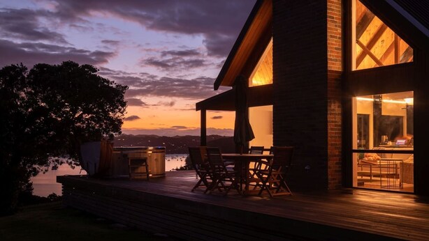 A modern house with a deck overlooking a body of water, featuring a dining area with chairs and a table, and a large window with a view of the landscape.