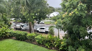 Ein Wohngebiet mit zwei weißen Lastwagen, die auf einer nassen Straße geparkt sind, umgeben von üppiger Vegetation und einem Haus im Hintergrund.