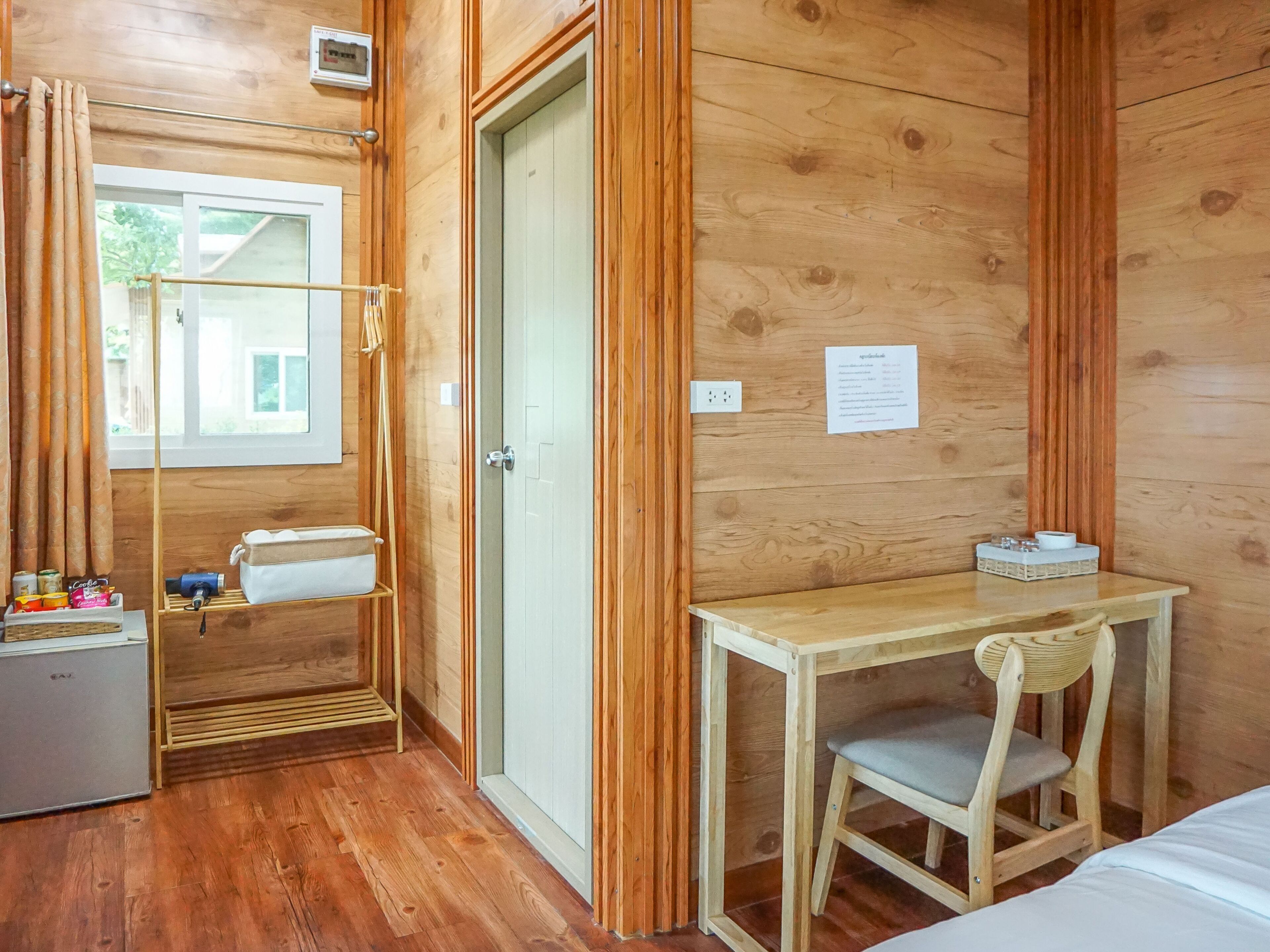 A wooden-paneled room with a bed, a desk with a chair, a small table, a shelf with items, and a window with curtains.