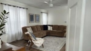 Una sala de estar con un sofá seccional marrón, un sillón blanco, una mesa de centro de madera y una alfombra estampada.