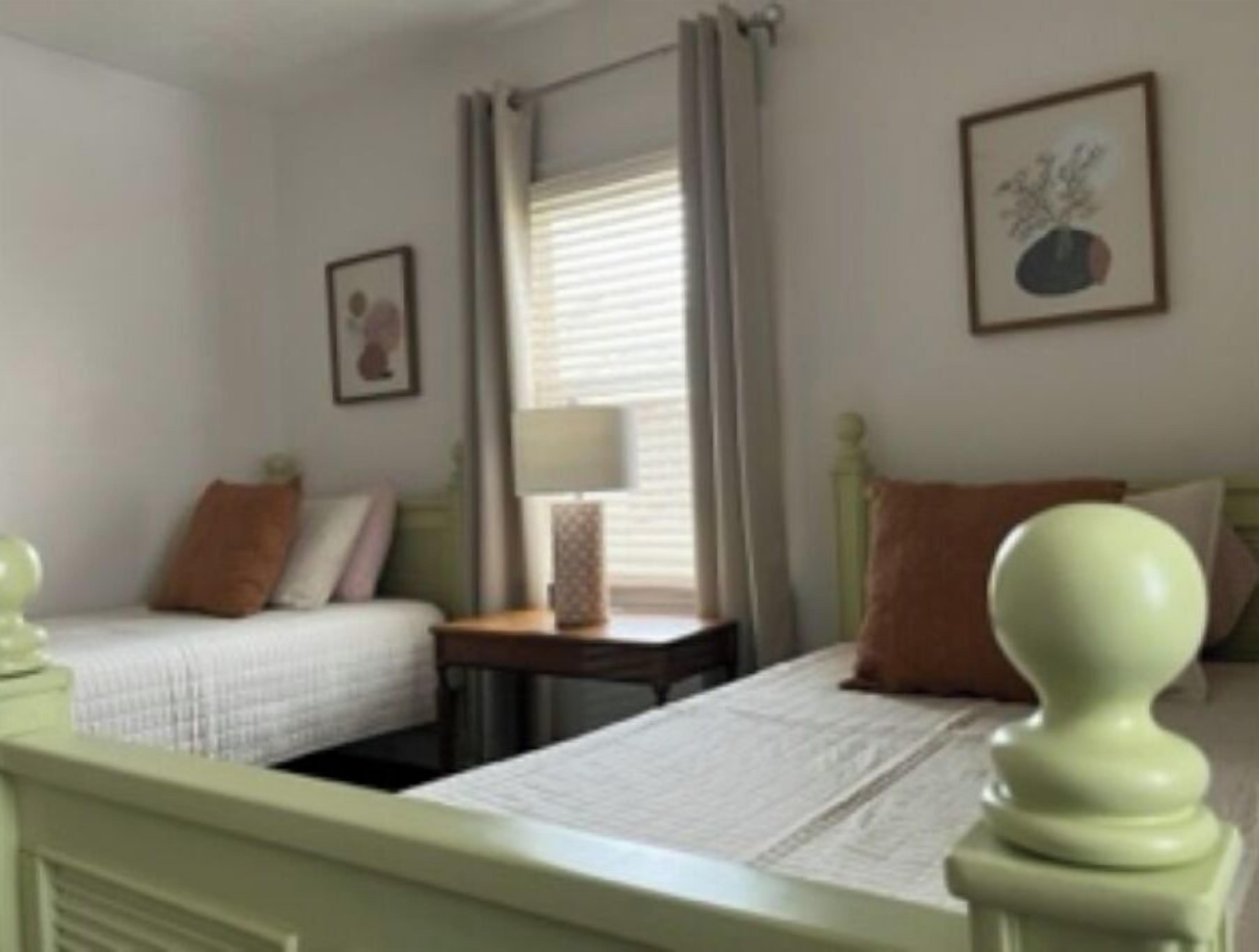 Une chambre avec deux lits simples, une table de chevet avec une lampe, une fenêtre avec des rideaux et un tableau encadré au mur.
