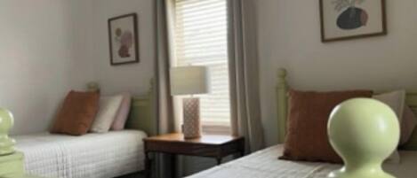Une chambre avec deux lits simples, une table de chevet avec une lampe, une fenêtre avec des rideaux et un tableau encadré au mur.