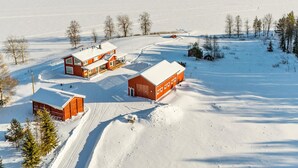 A snow-covered landscape with a large house and two smaller buildings, surrounded by trees and a frozen lake.