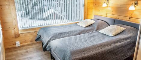 A cabin with two beds, a window with blinds, wooden walls, and a sloped ceiling.