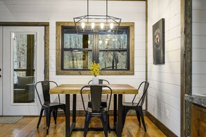 Een eetgedeelte met een houten tafel en vier stoelen, een vaas met gele bloemen en een groot raam met uitzicht op bomen.